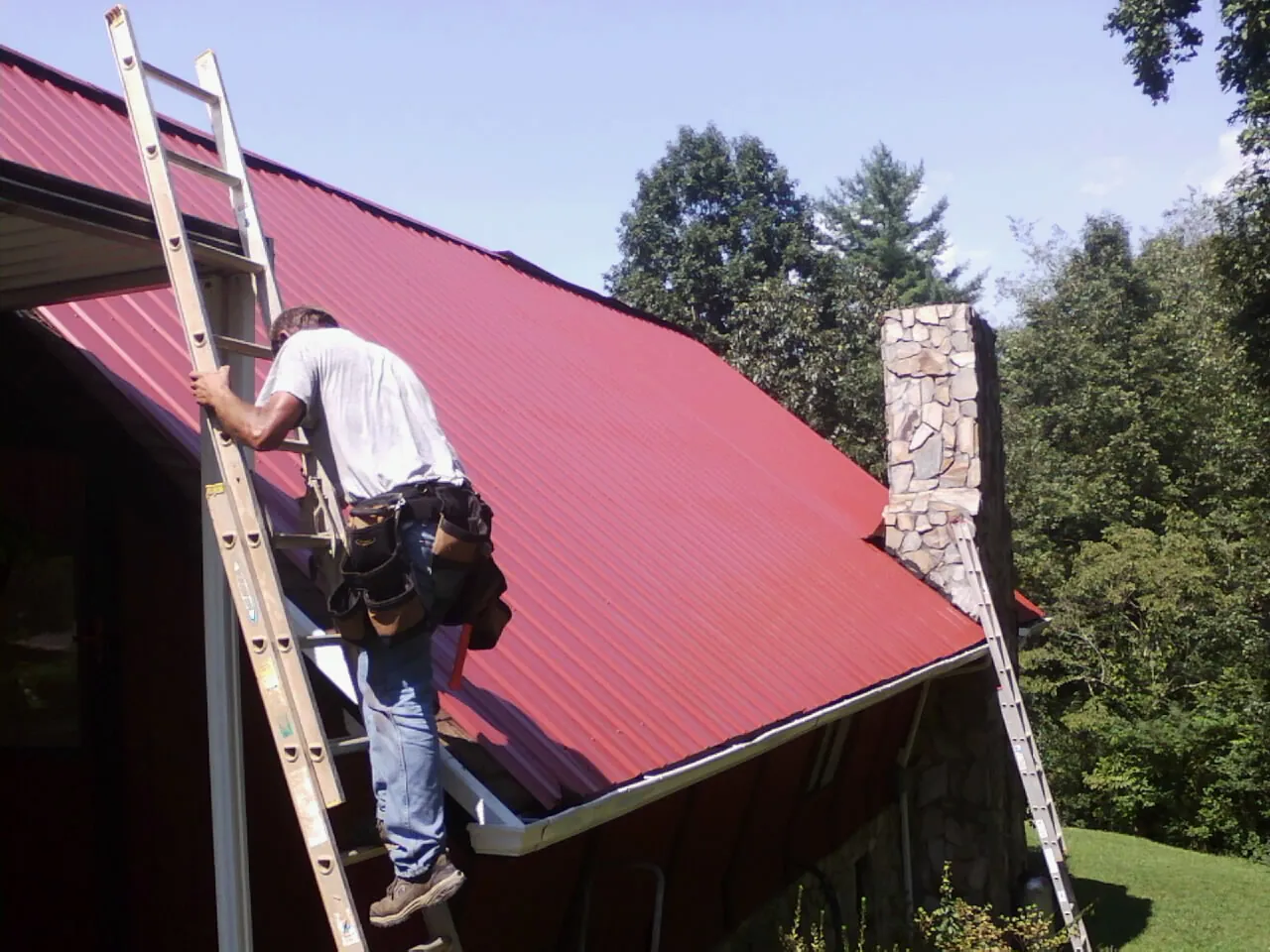Roofer on a ladder beside a red metal roof and stone chimney — Soffit & Fascia Repair by Roofing Brothers in Worthington